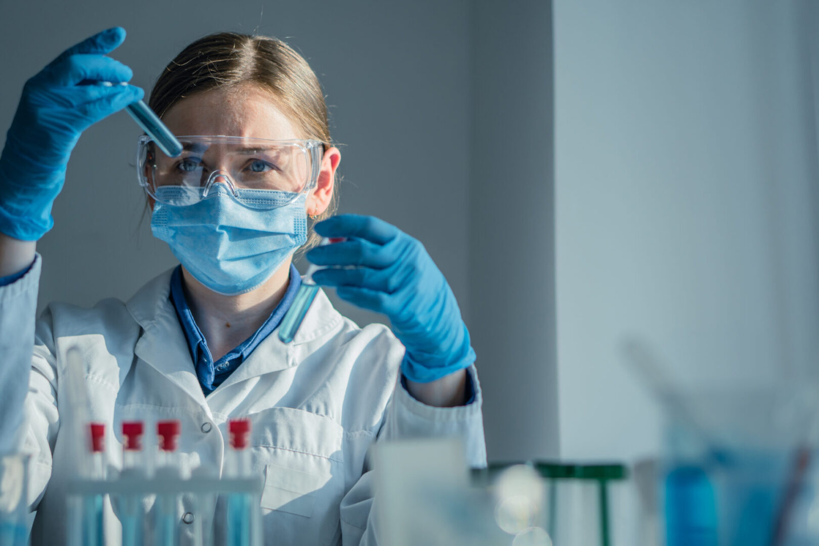 Scientist in a lab coat and mask examining a test tube with blue liquid.